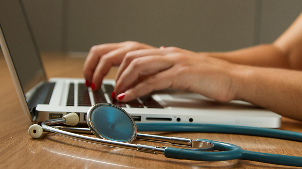 Hands typing on a laptop next to a stethescope