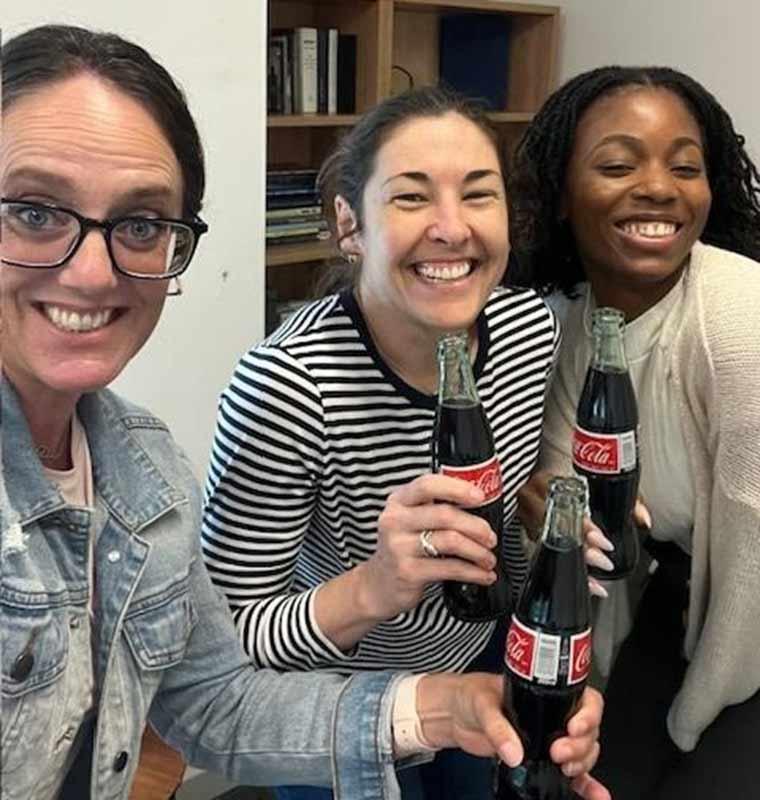 Three women clinking bottles of Coca-Cola together in celebration.