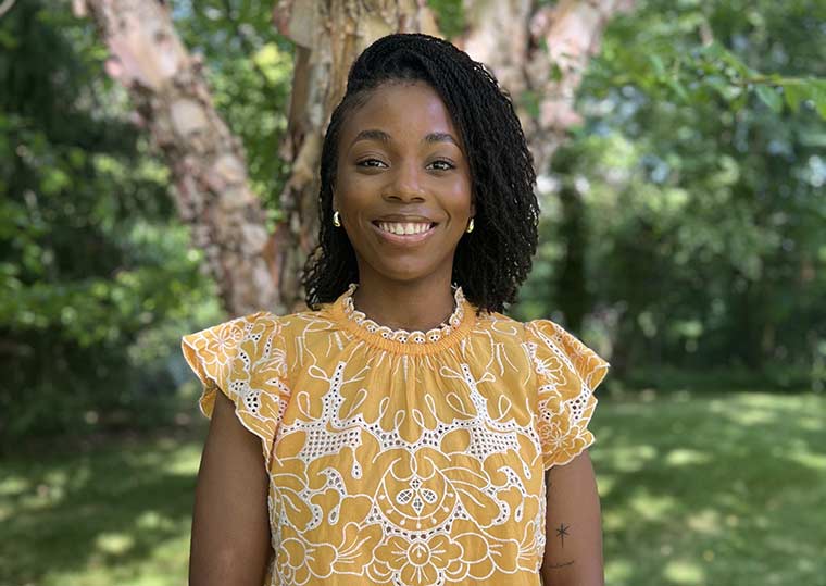 Picture of smiling woman with bright yellow shirt with white eyelet designs against a backdrop of a tree and grass