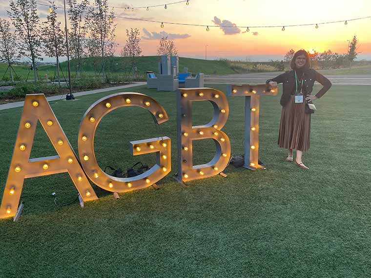 Woman next to large lit sign with letters ABGT with sunset in the background