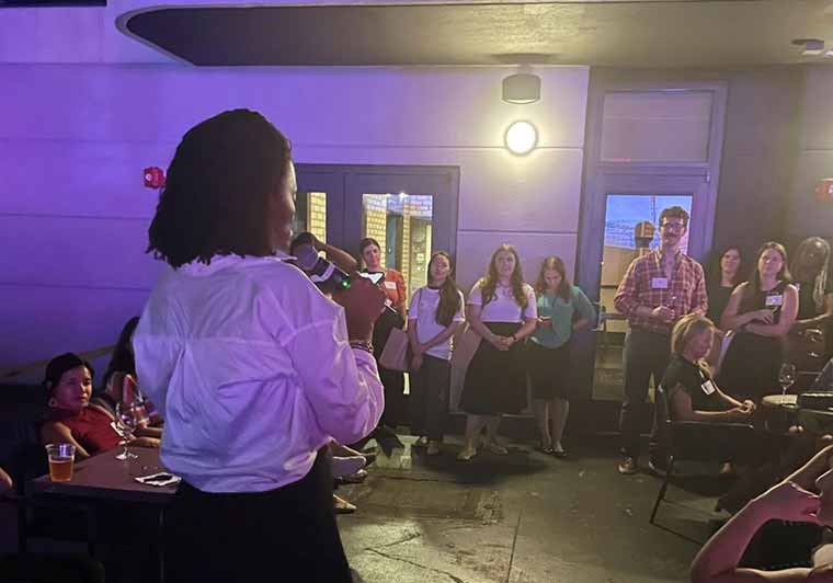 Woman speaking to a crowd at an evening event on a outdoor rooftop patio.